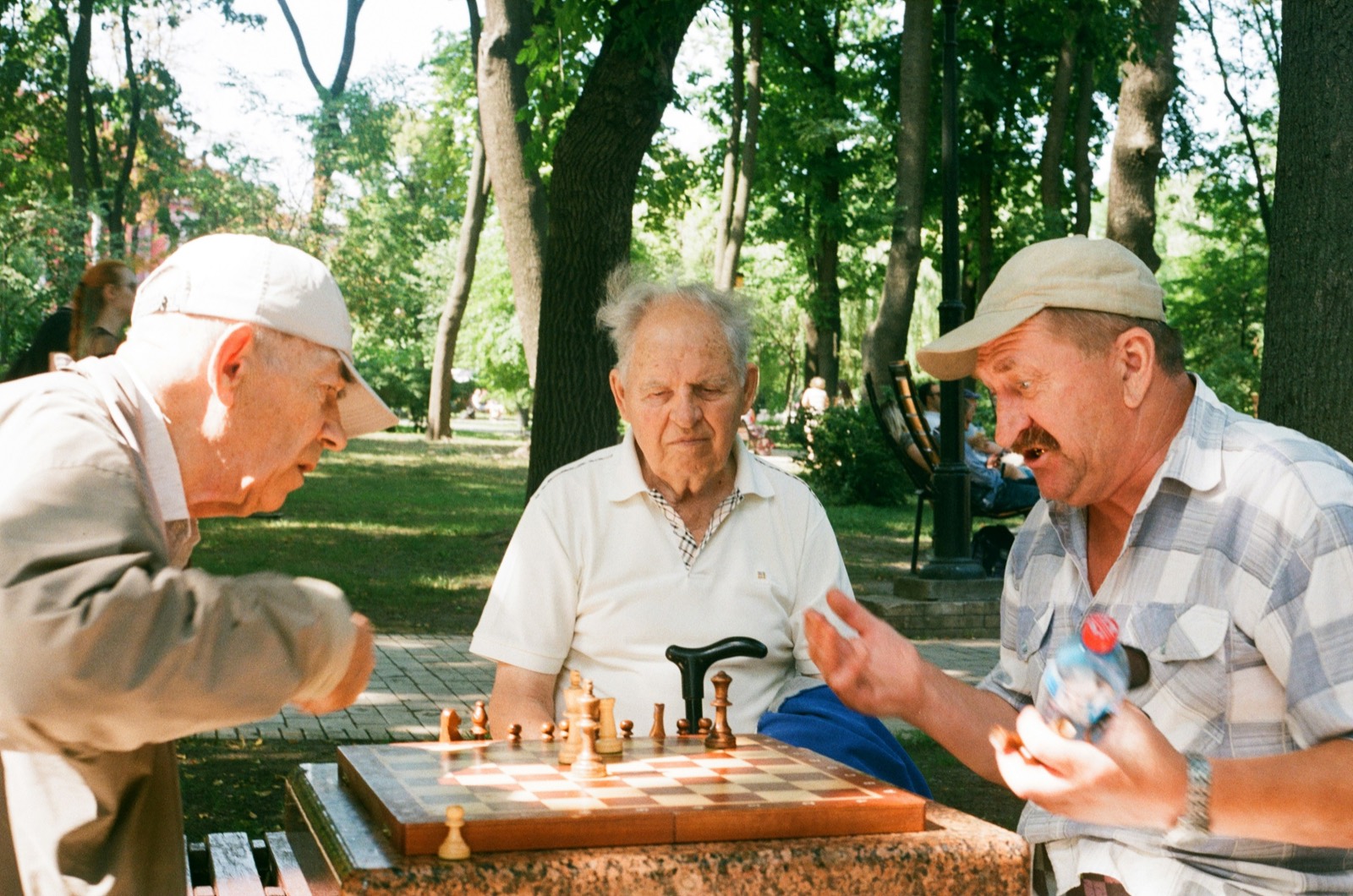 Three older men playing chess together at an outdoor table in a park